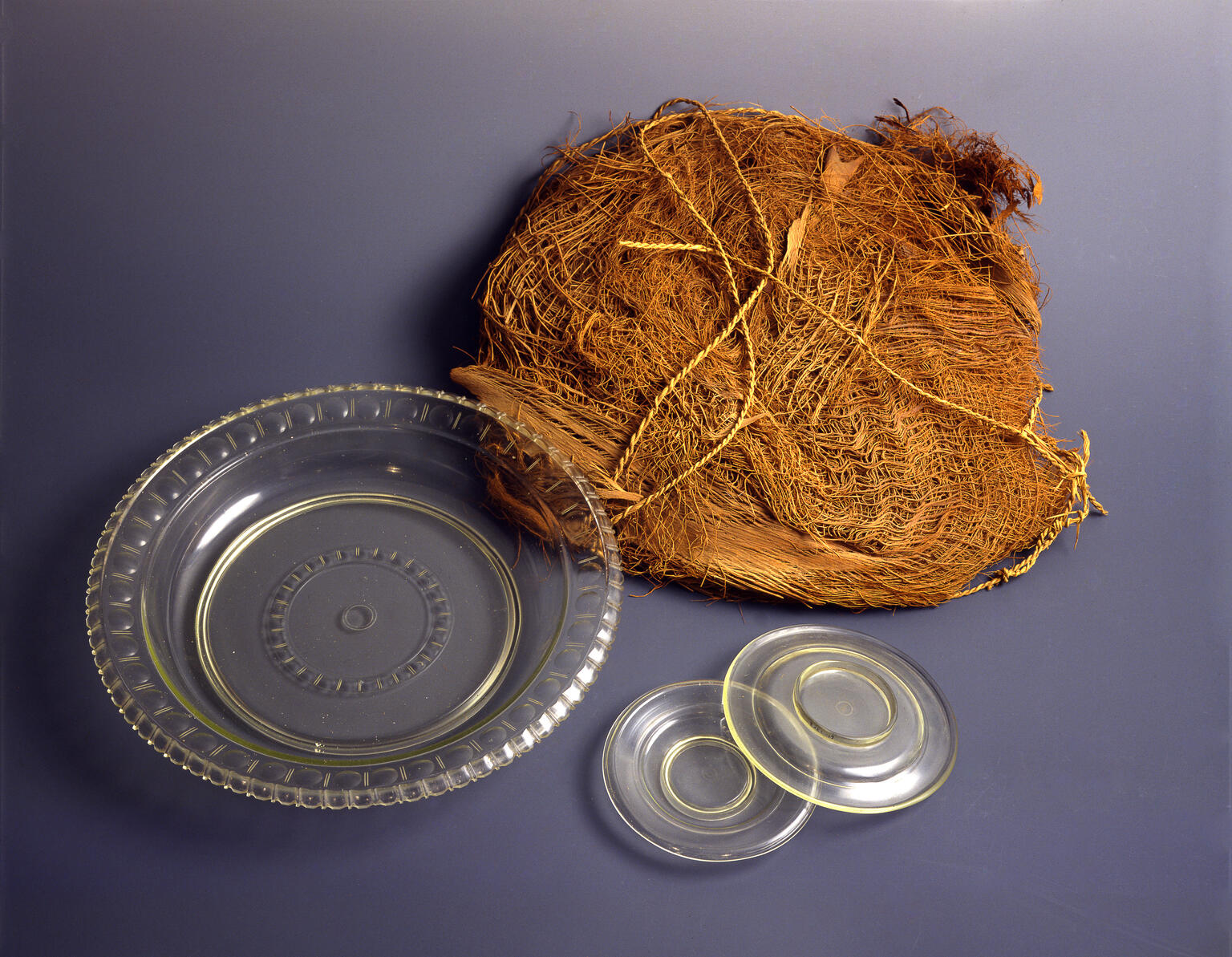 A bundle of palm fiber mat, a round clear glass dish, and two small clear glass dishes are arranged on a smooth, dark surface.