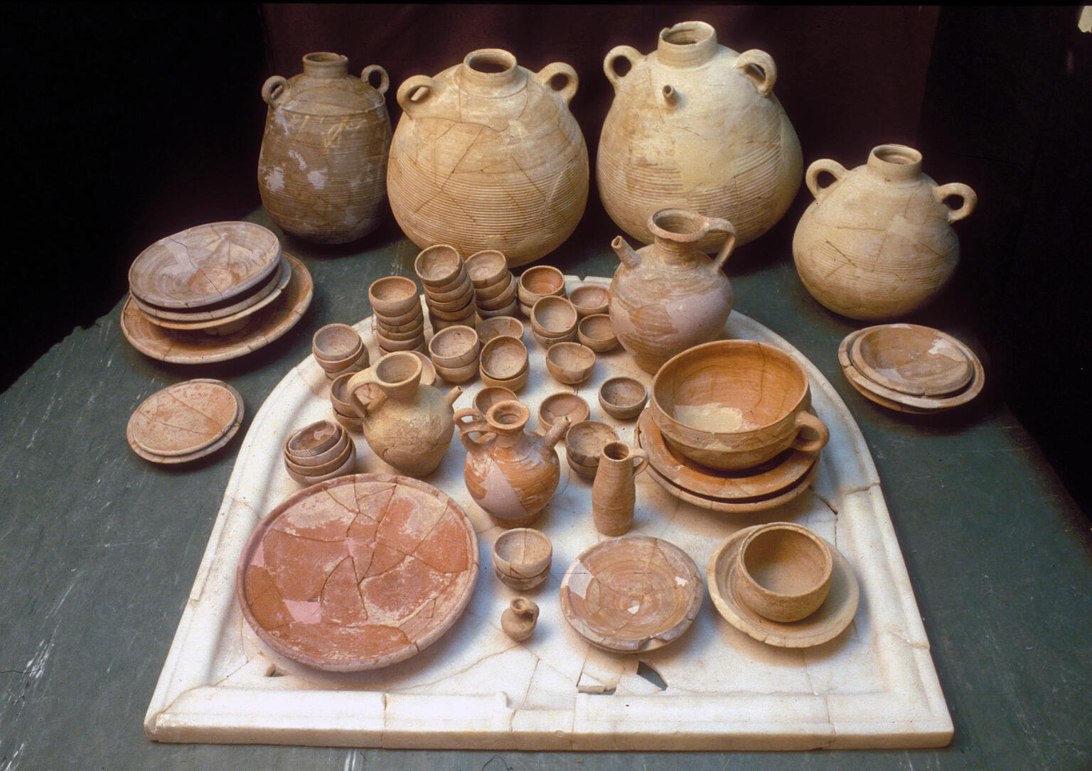 A collection of ancient ceramic jugs, bowls, plates, and cups in various sizes and earthy tones, arranged neatly on a stone surface with several large jugs in the background.