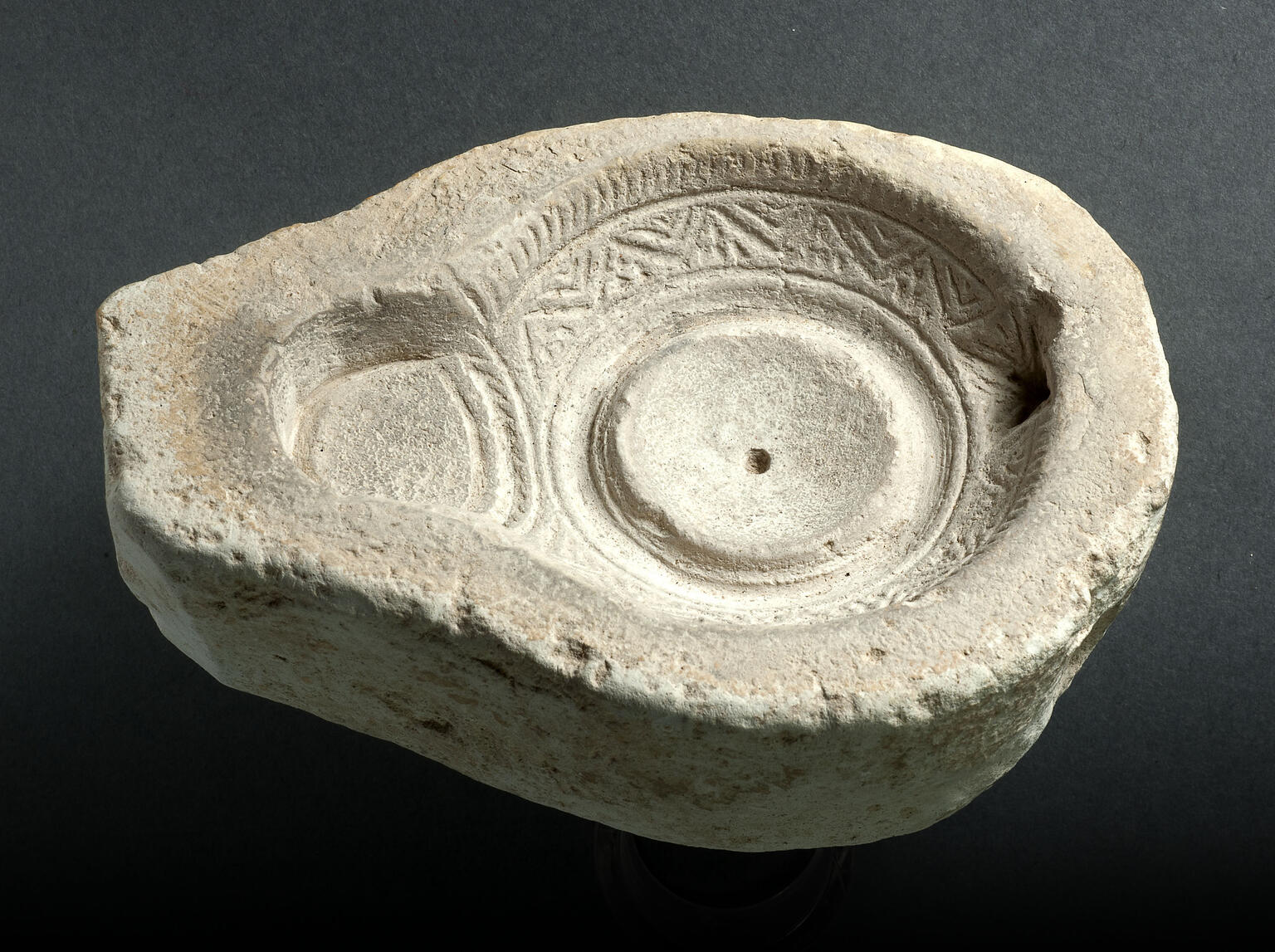 Ancient stone mold for an oil lamp with a circular reservoir, intricate geometric carvings around the rim, and a spout for the wick.