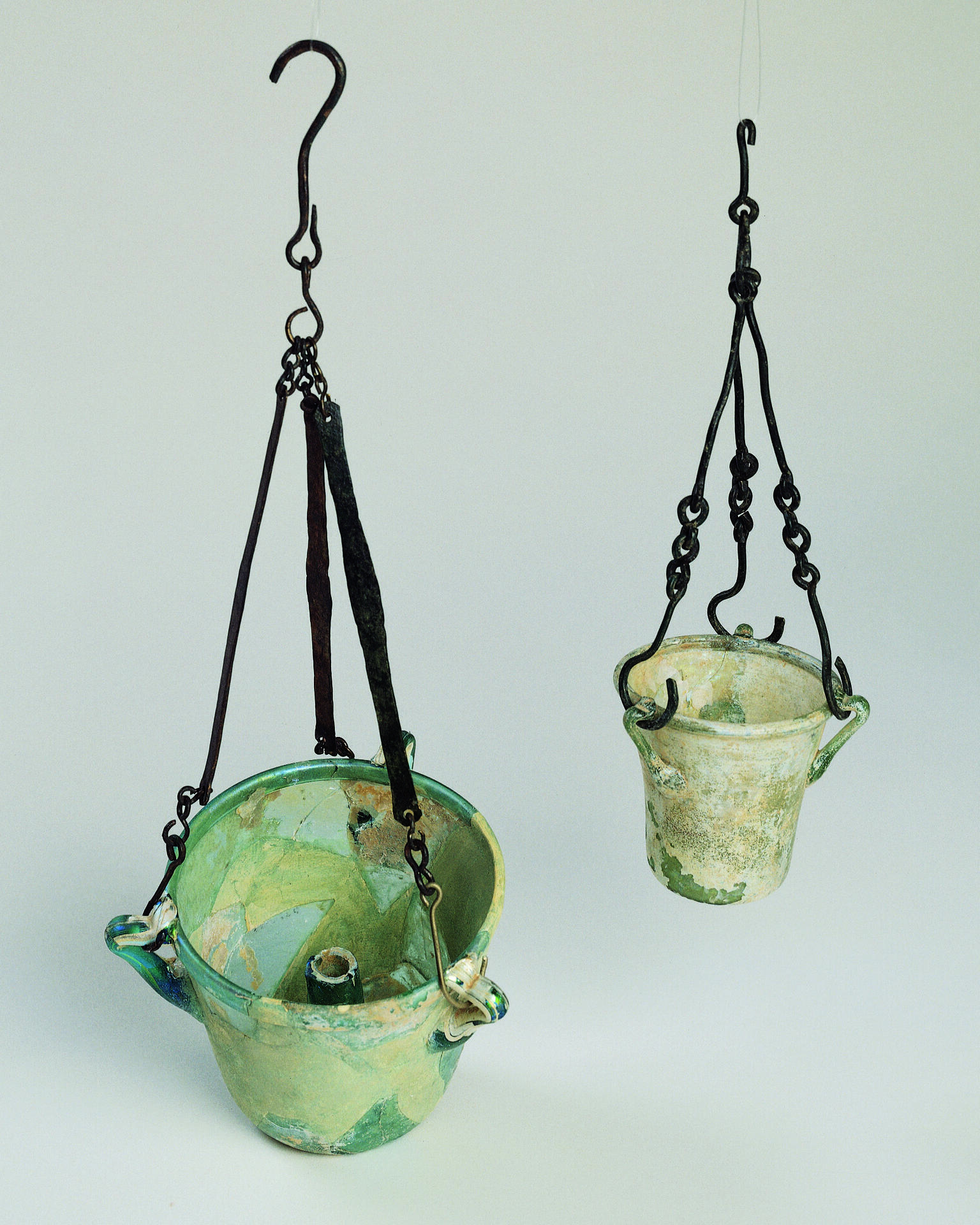 Two ancient greenish, bucket-shaped glass lamps hanging against a plain background.