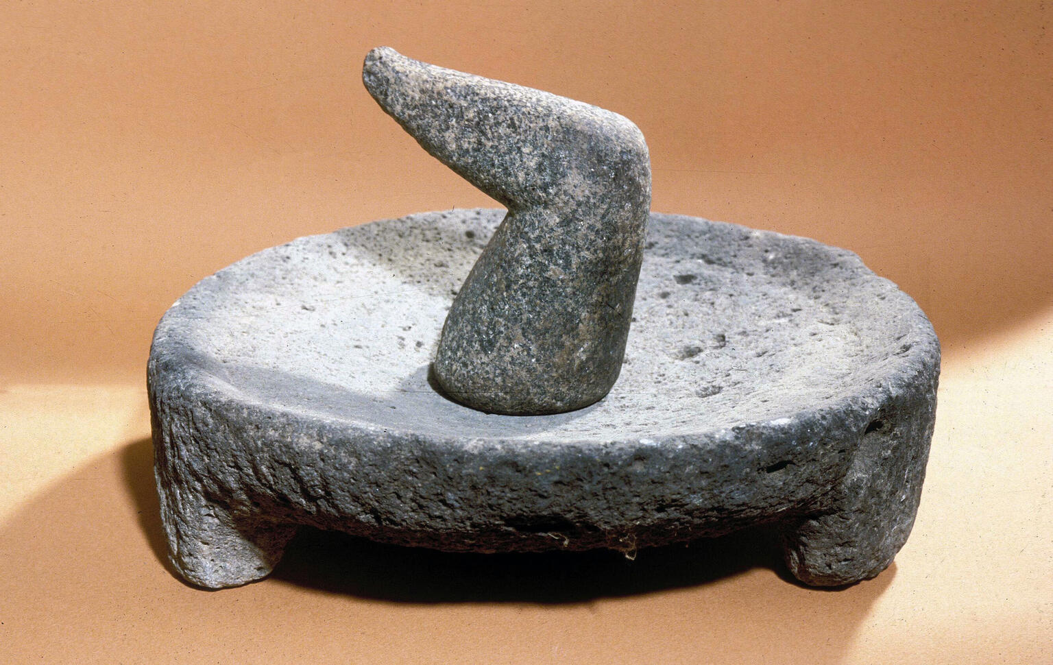 A grinder with a flat, round base and a hand-held cylindrical stone. The objects are gray and sit on a light brown surface.