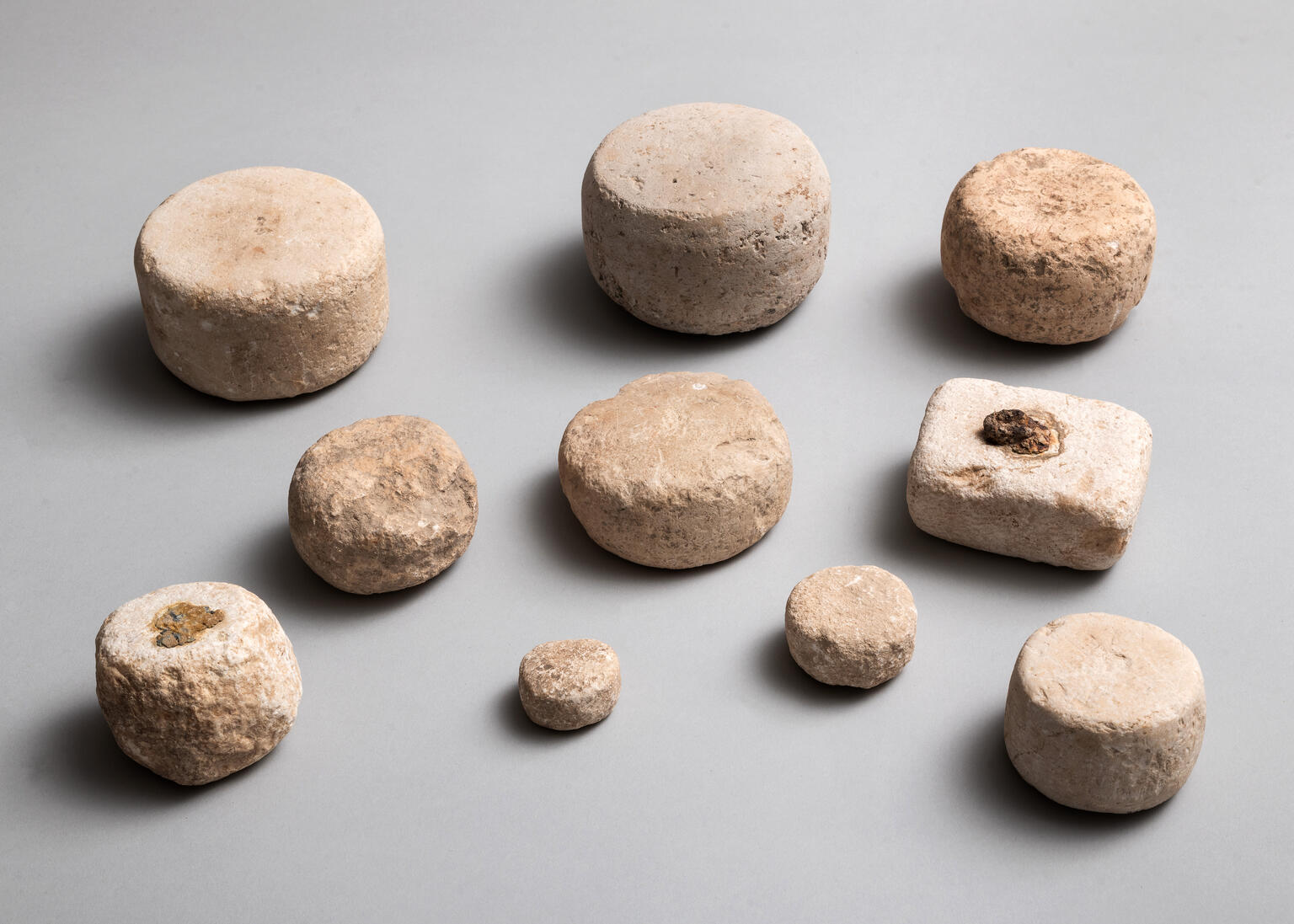 Ten ancient stone weights of various sizes and shapes, mostly cylindrical and one roughly rectangular, arranged on a plain gray background. The stones appear worn and textured, with earthy beige tones.