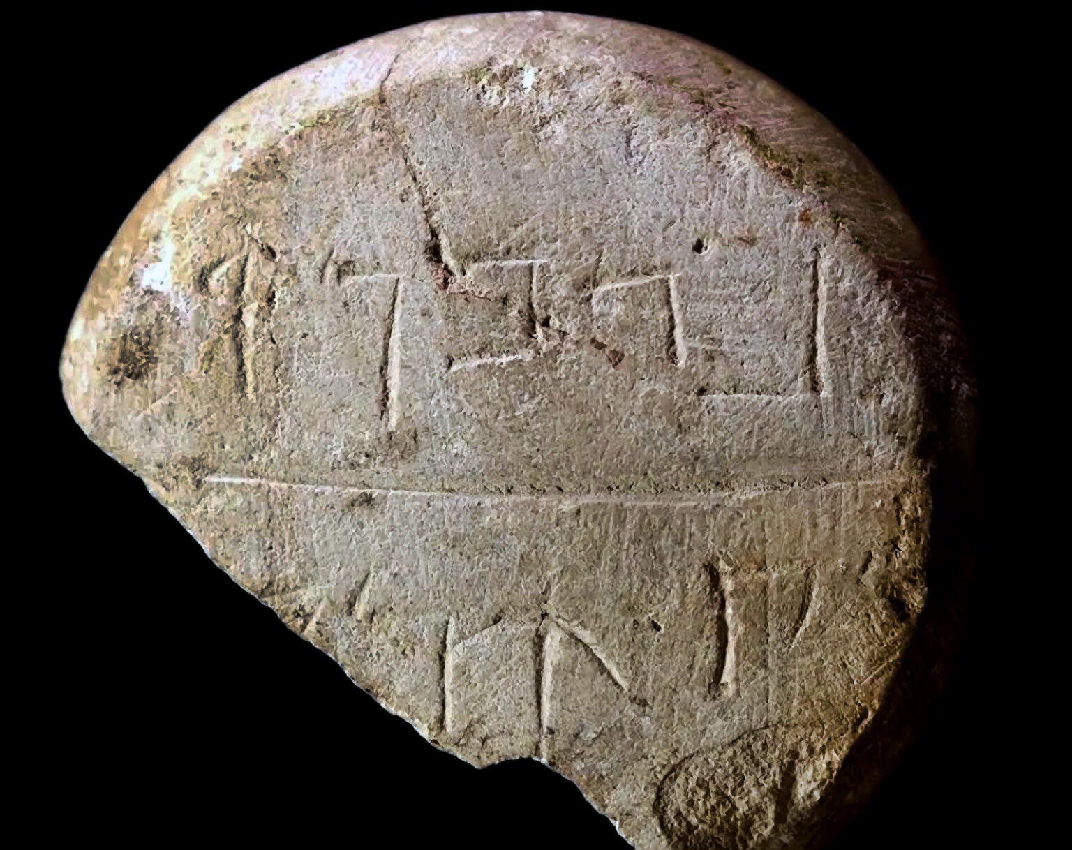 A broken, round stone with ancient script carved into its rough surface, featuring two visible lines of inscriptions, against a black background.