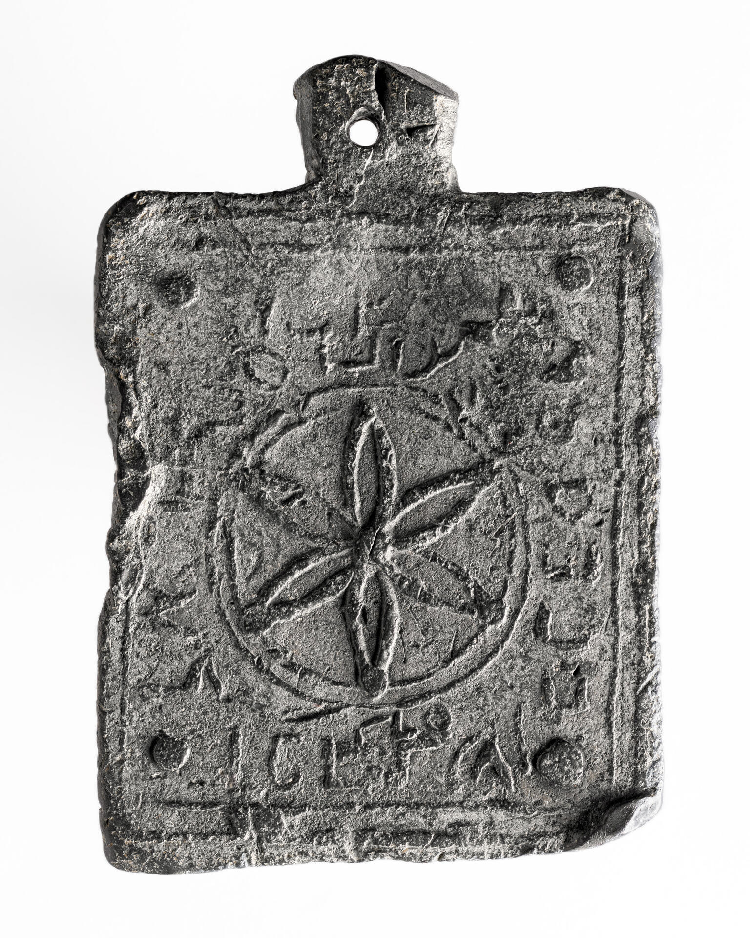 A rectangular, weathered metal object with a central flower-like geometric pattern inside a circle, surrounded by worn Hebrew inscriptions, with a hole at the top for hanging.