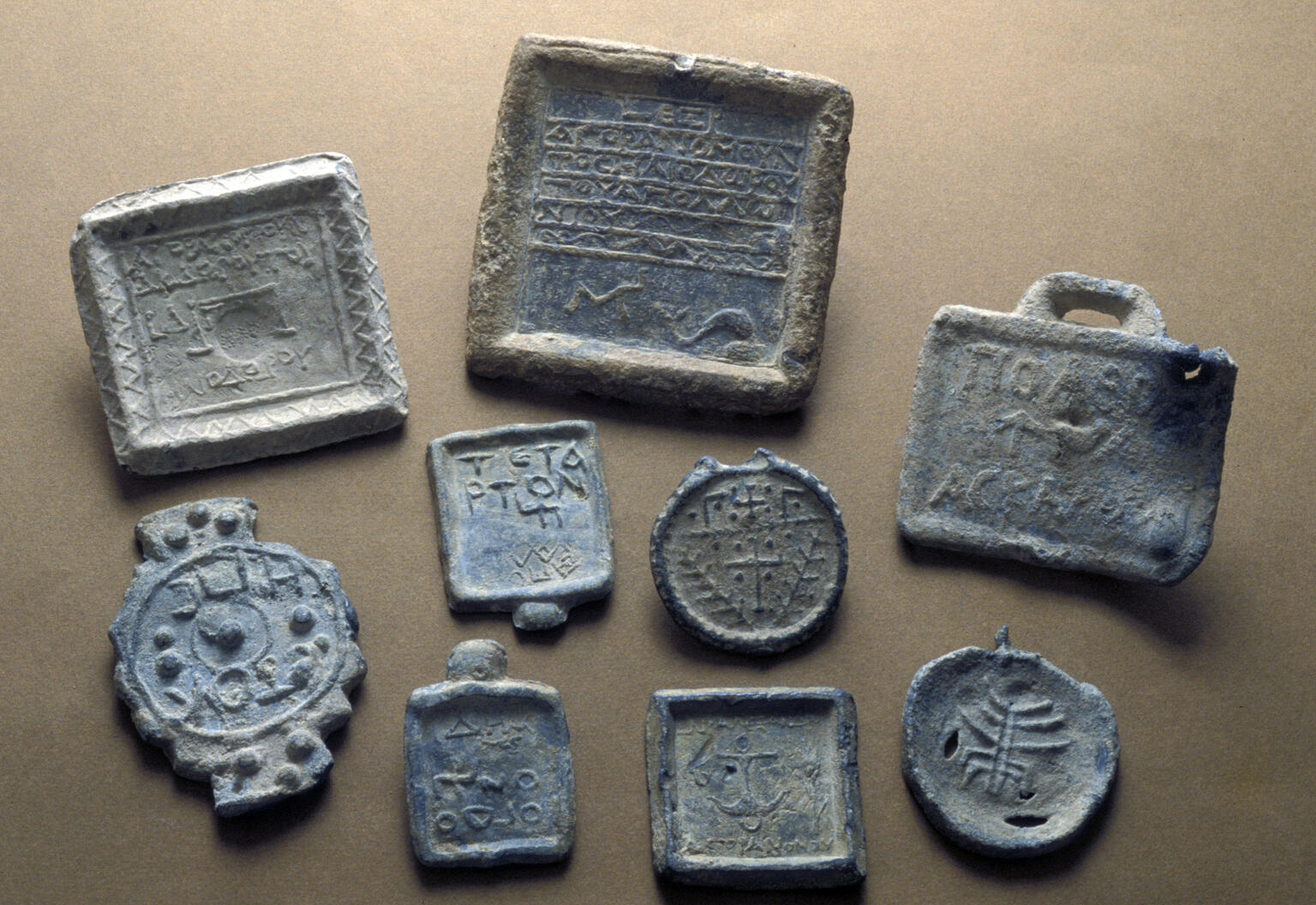 Nine ancient lead weights with various shapes, engravings, and inscriptions are arranged on a flat, brown surface. Some feature symbols, while others display text.