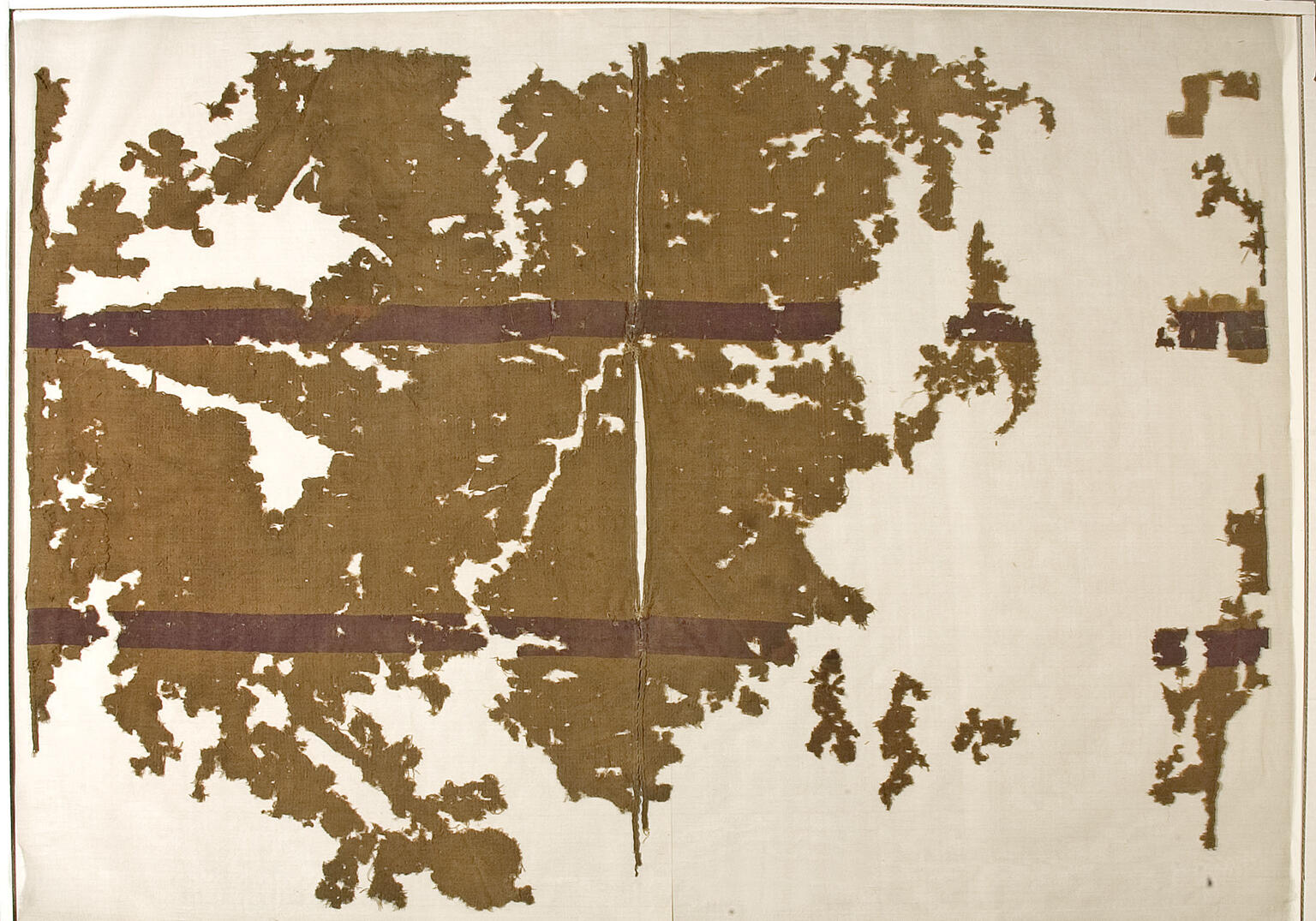 A brownish, ancient textile fragment with dark red horizontal bands on a cream background, showing heavy wear and missing sections.