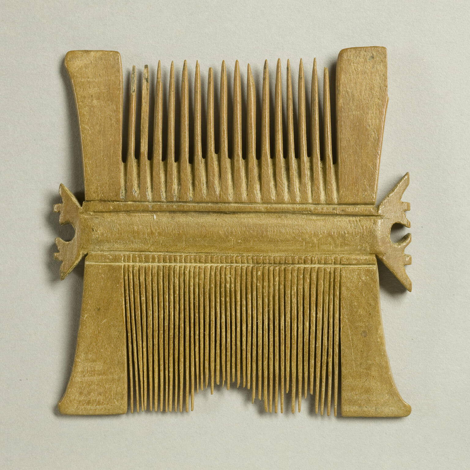 A double-sided, ancient wooden comb with wide teeth on one side and fine teeth on the other, showing some broken and uneven teeth.