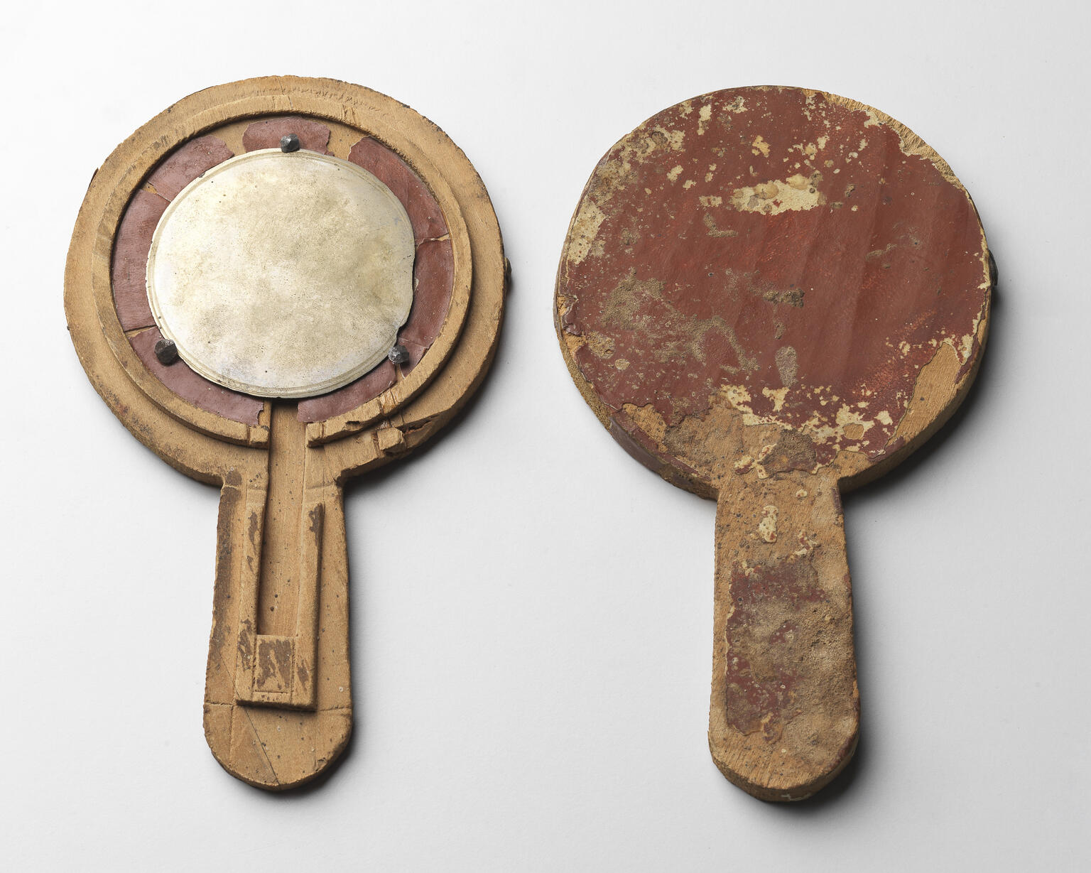 Two ancient wooden hand mirrors with round shapes and handles. The mirror on the left has a reflective circular metal surface; both show signs of heavy wear, scratches, and peeling surfaces.