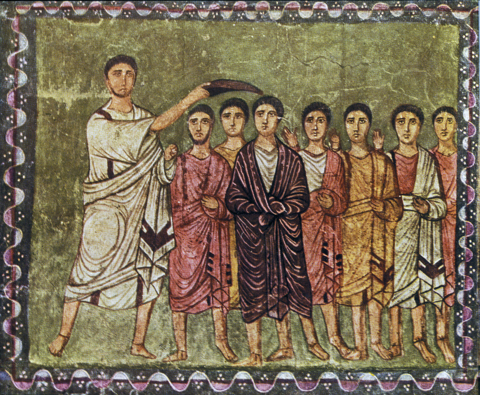 A painting shows a figure in a white robe anointing the head of another figure standing among several others, all dressed in robes, against a background with a decorated border.