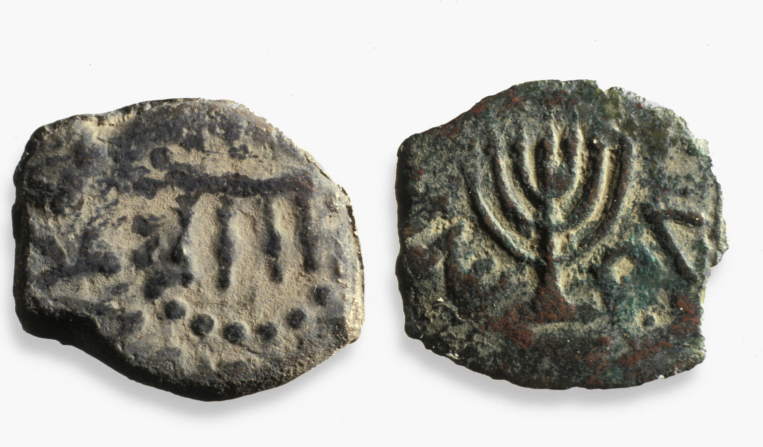 Two sides of an ancient bronze coin: one shows a four-legged table surrounded by dots, and the other features a seven-branched menorah. Both sides have faint inscriptions. The coin appears worn and weathered.