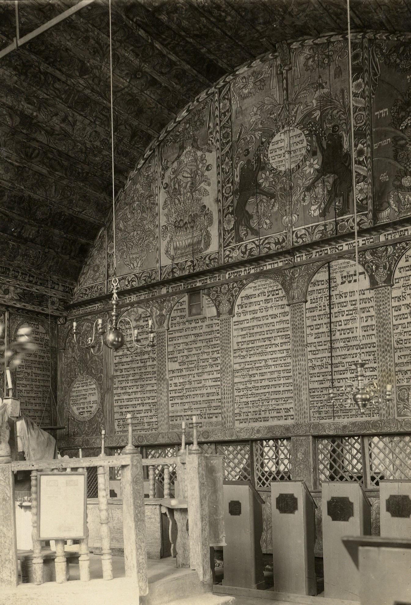 Interior of building of back wall with decorative images on top, Hebrew text in the middle, and lattice below, platform with railing in left foreground, and several podiums along wall.