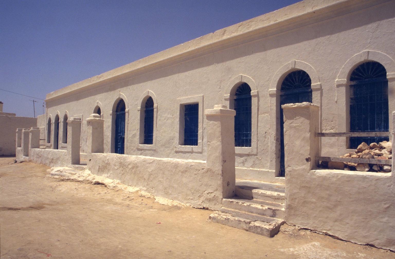 Exterior photograph of stone building with arched windows, wraparound patio with stone columns, and dirt ground.