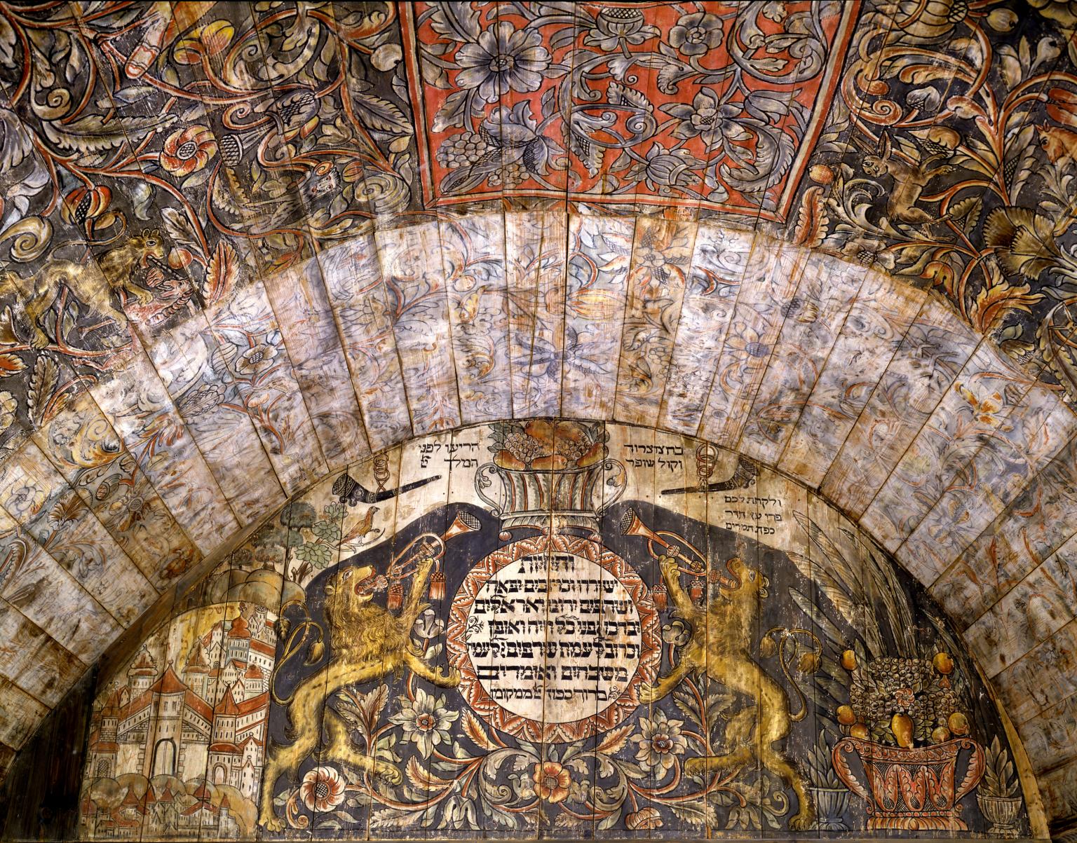 Close up photograph of wall and ceiling with painting of lions on either side of Hebrew text, city on left side, cornucopia on right side, and floral and plant designs on ceiling. 