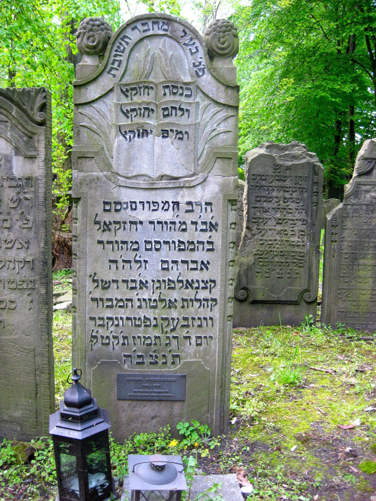 Tombstone of relief of open books, Hebrew inscriptions, and two lanterns on ground in front. 
