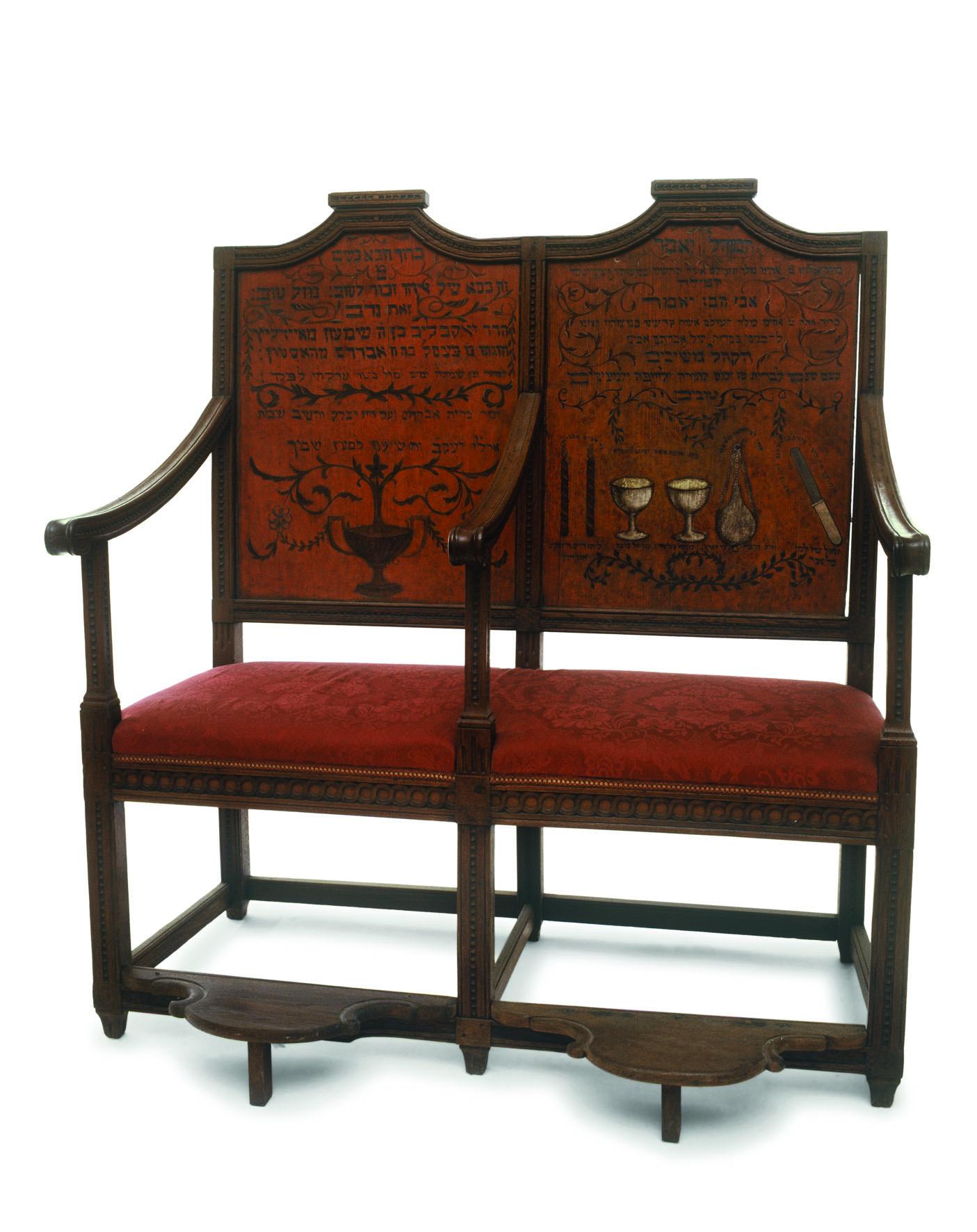Two-seater bench with central armrest, decorated on the seat backs with ritual objects and Hebrew text.