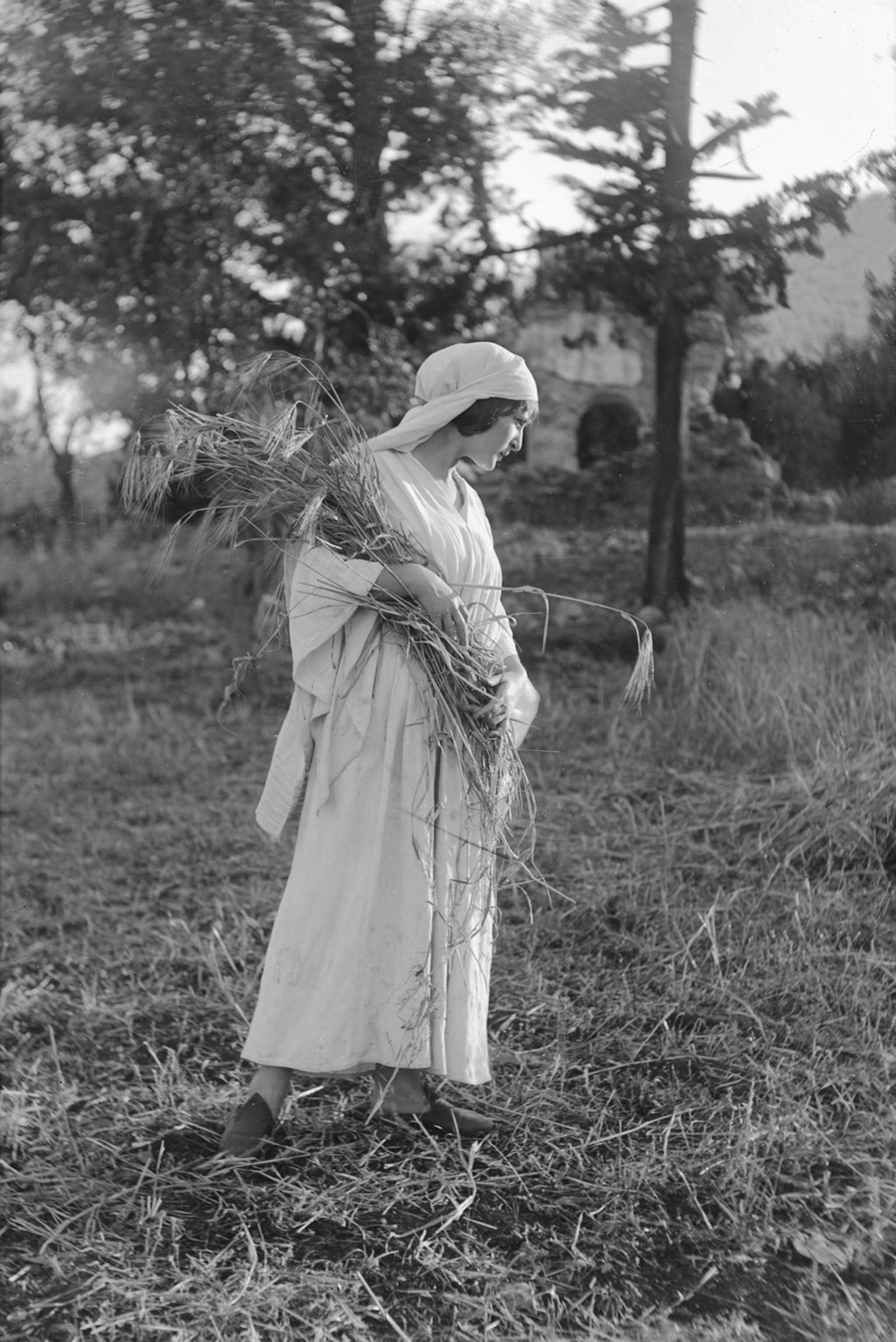Photograph of woman wearing white head scarf holding a bouquet and gazing downward in a field.