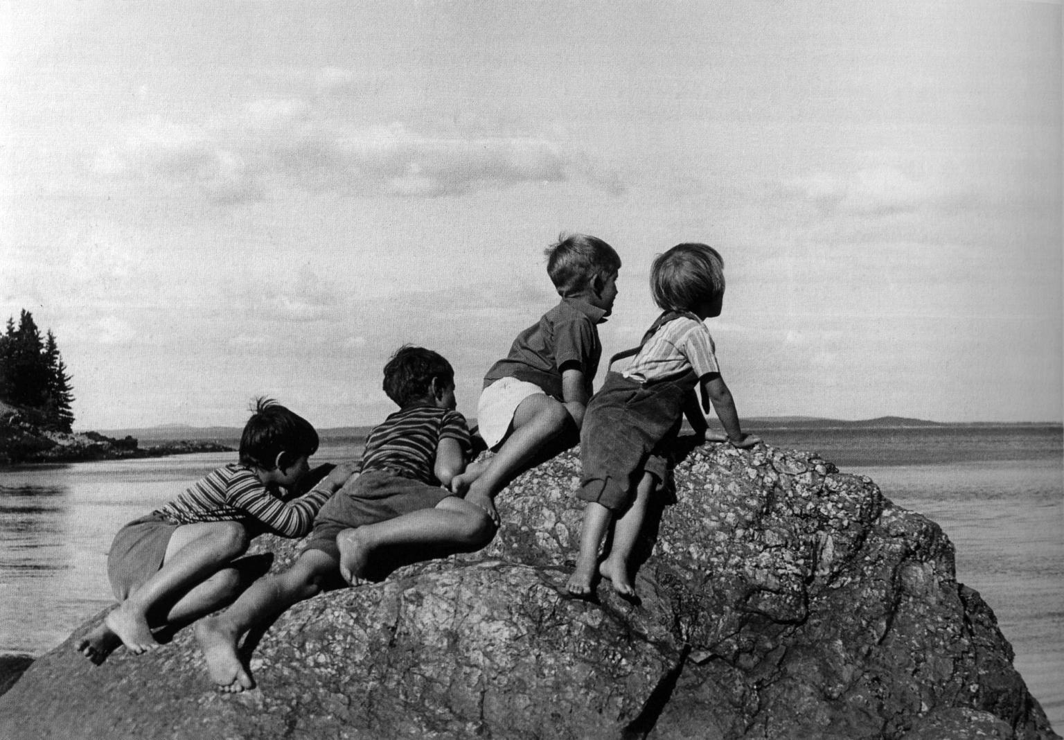 Photograph of four boys perched on a rock overlooking water and facing away from viewer.