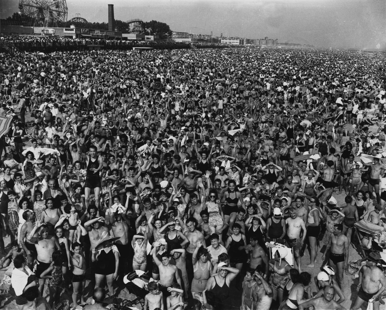 Aerial photograph of hundreds of people crowded together on beach with amusement park in background.