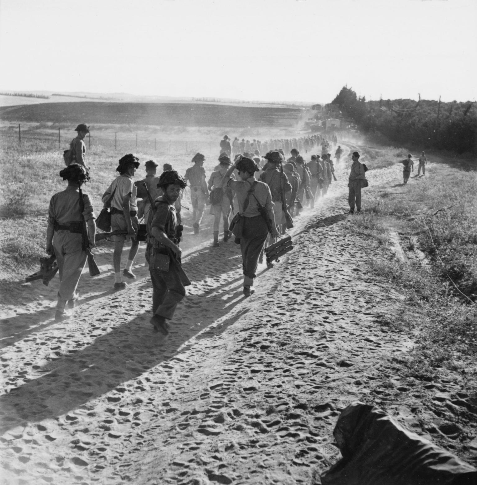 Photograph of troops marching away from the viewer on a sandy trail.