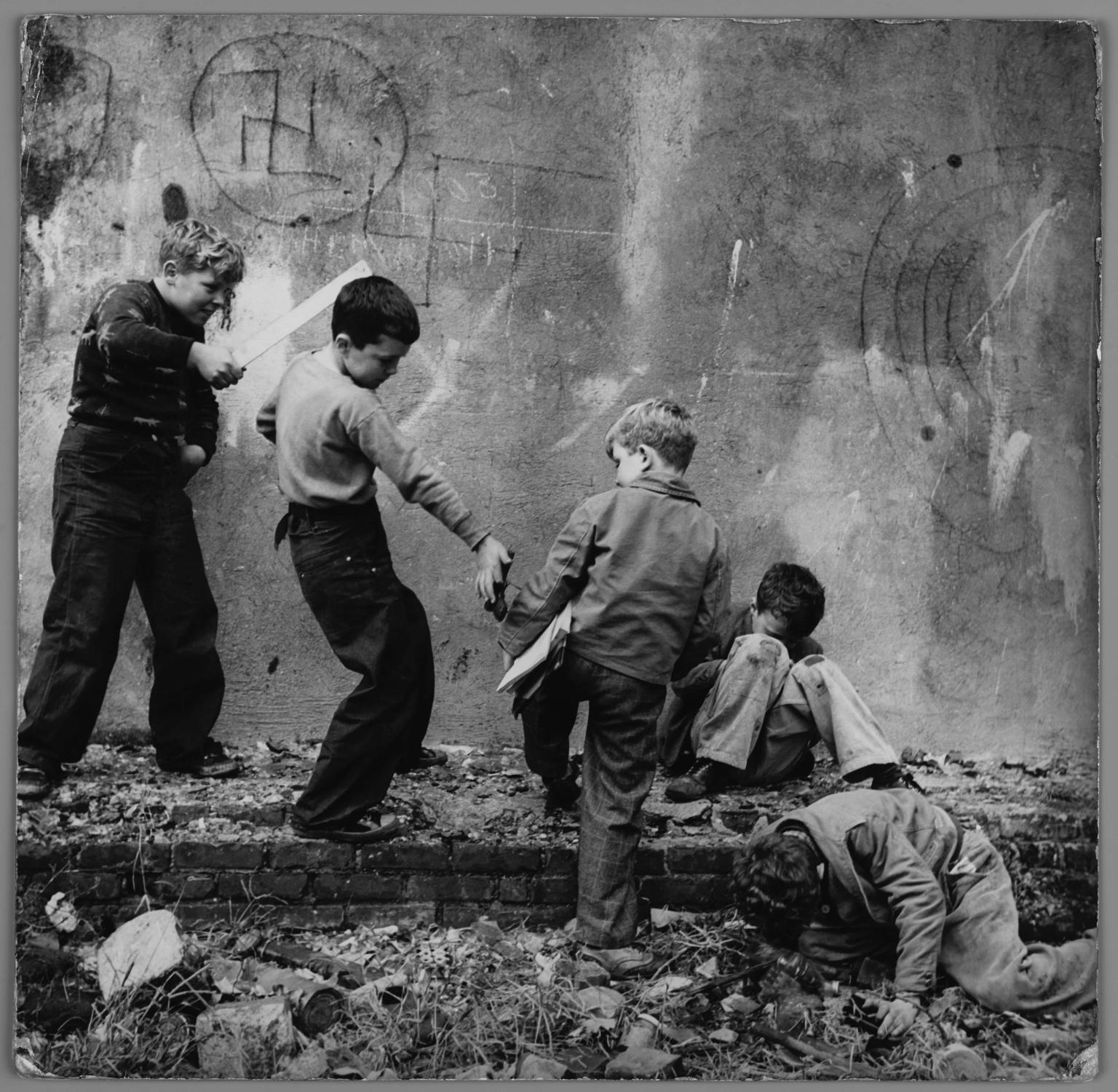 Photograph of five young boys standing and sitting in various poses on crumbling foundation of a wall with swastika drawn on it. One boy points a flat wooden stick at another boy, who is giving a metal object to a seated boy. 