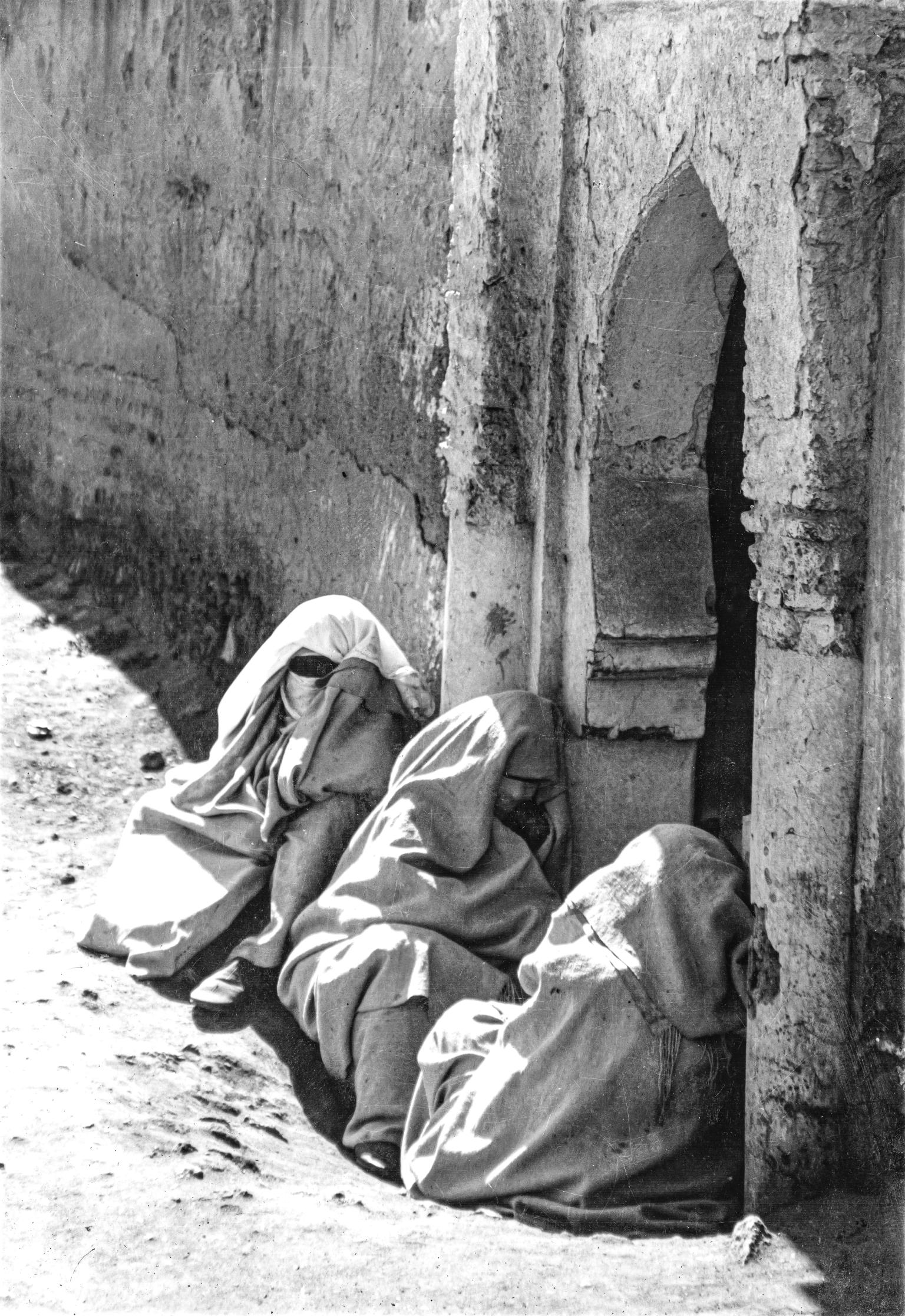 Photograph of three women seated on the floor next to building wearing hooded robes and veils and angled away from the viewer.
