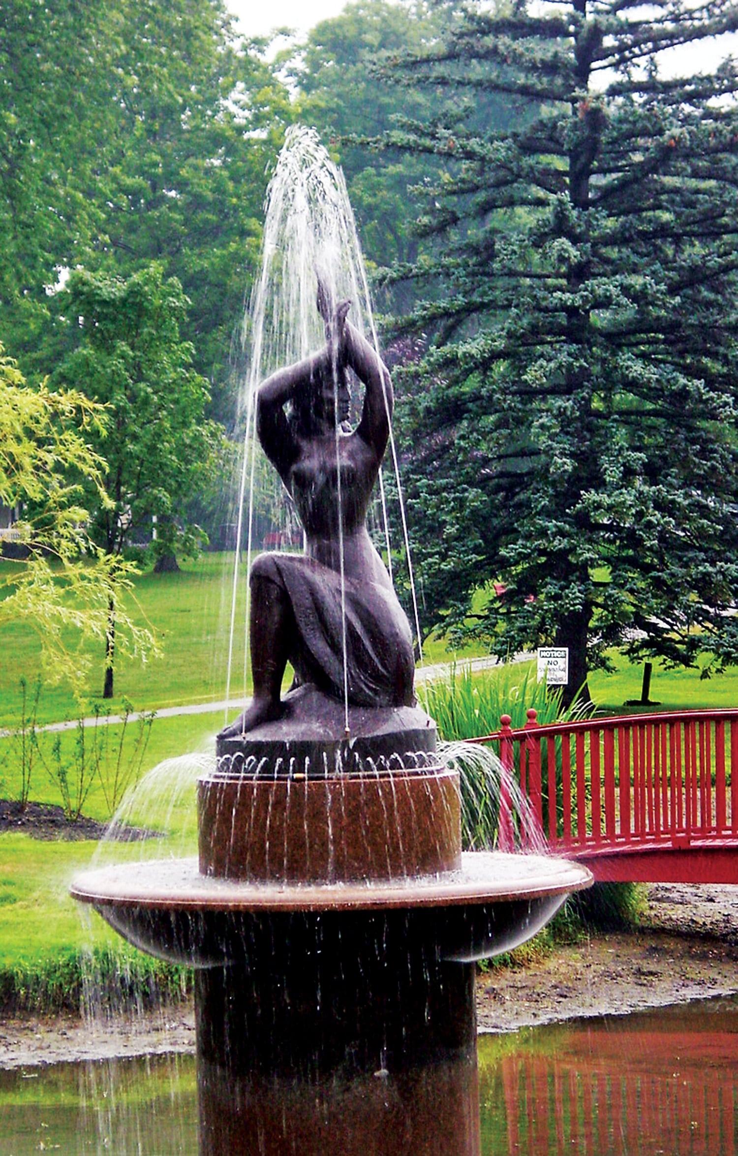 Bronze sculpture and fountain in the shape of a partially nude woman, resting on one knee and raising her arms above her head, set in a park with trees and a bridge in the background. 