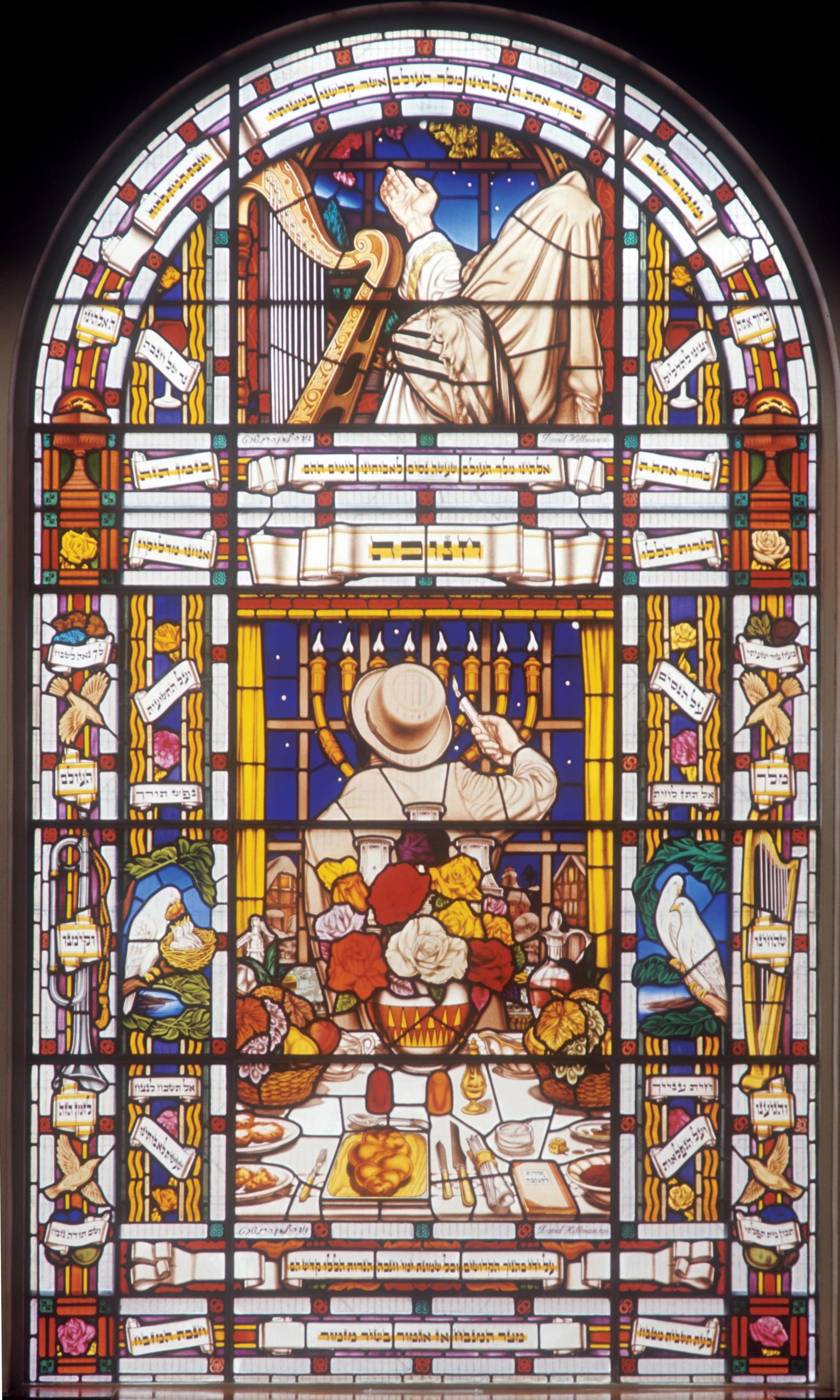 Stained glass window featuring a man in the center facing away from viewer lighting a menorah behind a table set for Shabbat, a rabbi above him next to a harp, and biblical images and Hebrew words, flowers, birds, and other objects.