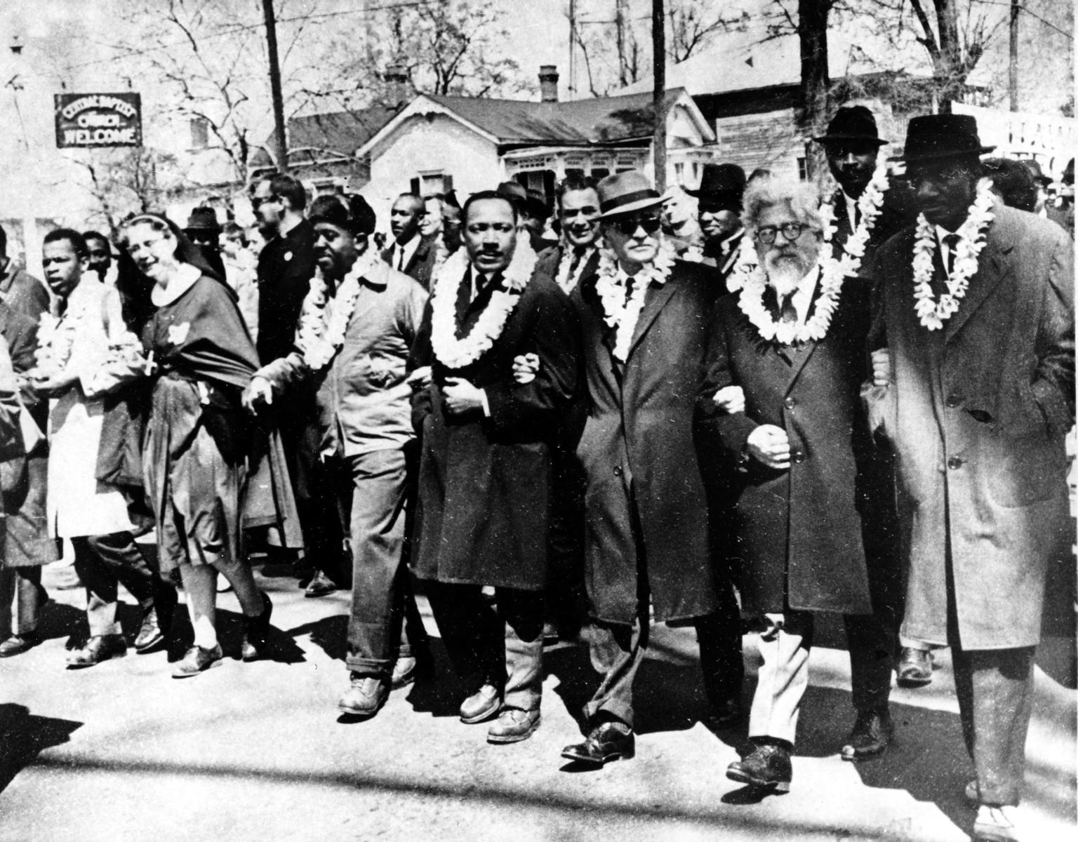 Photograph of men marching down the street with arms linked wearing white double carnation leis. 