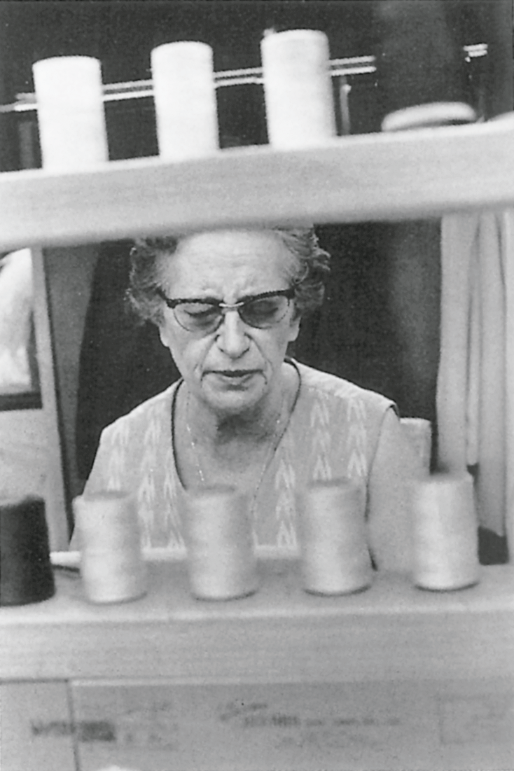 Photograph of elderly woman with glasses framed by spools of thread. 