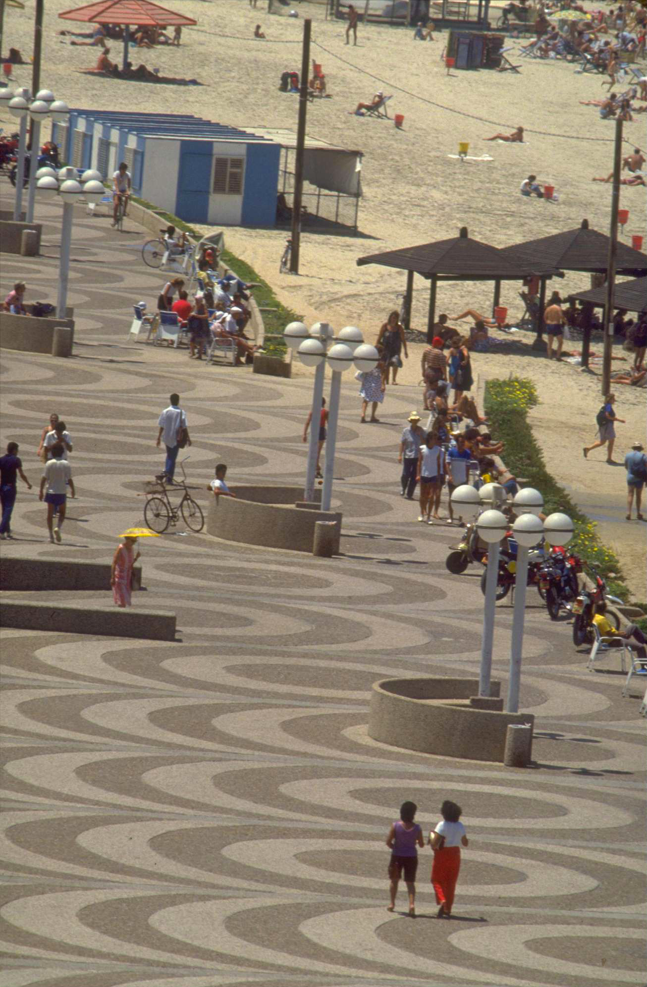 Photograph featuring a promenade alongside a beach with people walking and sitting, with several sets of lightposts and circular benches.