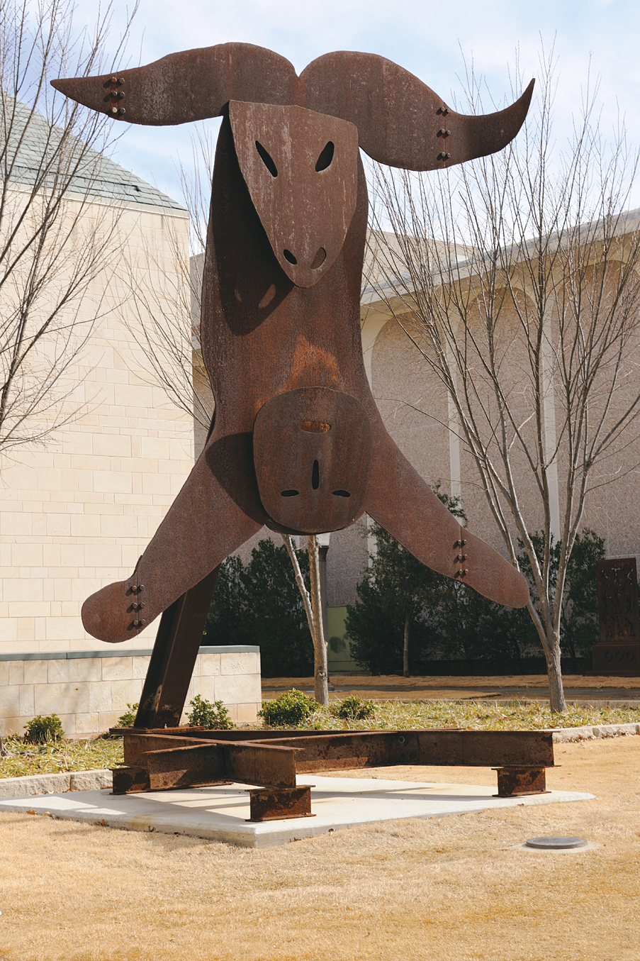 Steel sculpture on steel frame outside of building. Top of sculpture is the head of a sheep with horns and bottom of sculpture is stylized head and arms of a human figure.