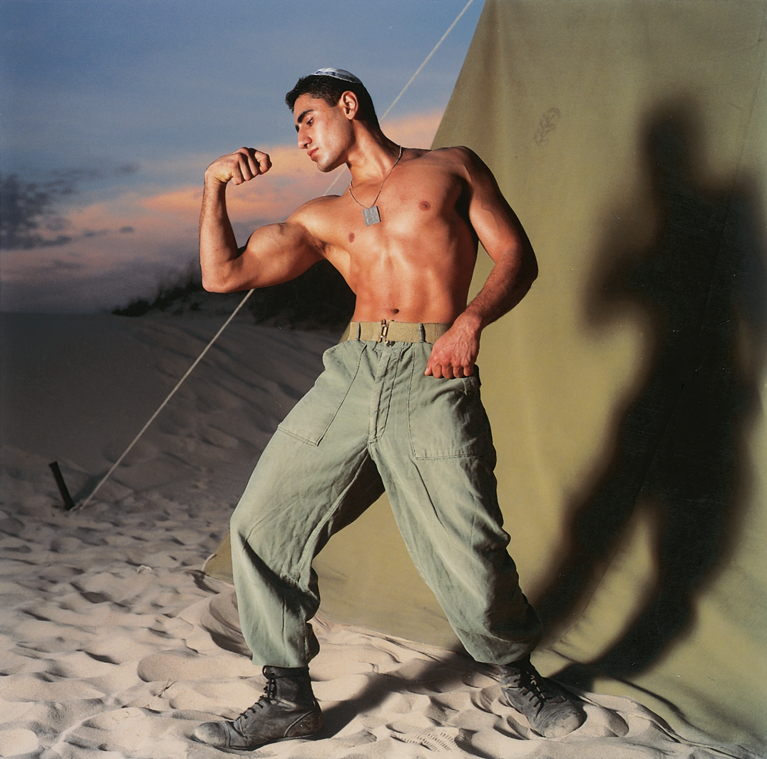 Photograph of man in kippah with shirt off flexing his bicep outside of tent. 