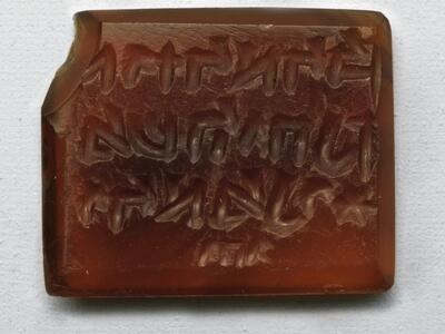 Photo of a rectangular red stone block with a beveled edge and chipped corner, inscribed with reversed Hebrew writing in three lines followed by a small word.