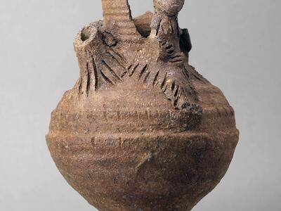 Earthenware vessel with a round body and flared base, with sculpted elements, some broken off, at the top, including a humanoid figure.
