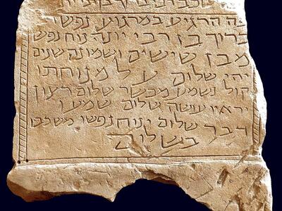 Photo of a broken, jagged stone slab with a Hebrew inscription, including a separated heading, decorative borders, and a simple candelabrum.