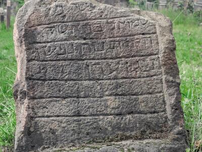 Photo of a weathered, broken gravestone in a cemetery, with seven delineated spaces, four of which contain Hebrew inscriptions.