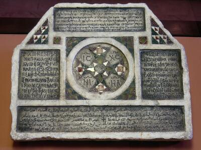 Photo of a hexagonal stone slab engraved with thickly-borded inscriptions in Latin, Hebrew, and Arabic, among which are geometric decorations, including, centrally, a circular pattern with four Latin letter pairs.