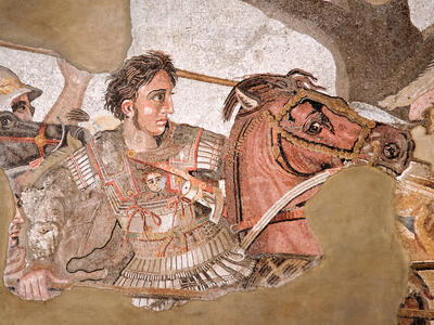 Ancient mosaic depicting a warrior in ornate armor, holding a spear, riding a horse into battle. The warrior has wavy hair and a determined expression and is surrounded by other soldiers. The background is partially damaged.