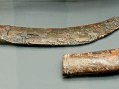 An ancient, curved metal dagger and scabbard displayed side by side on a flat surface. The dagger has a corroded blade and worn surface, and a hilt with three rounded knobs.