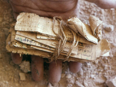 Close-up of a hand holding an ancient, rolled bundle of papyrus tied with string, resting on a dusty surface with small rocks or pieces of debris scattered around.