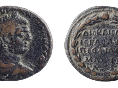 Ancient coin with a profile of an emperor on one side and inscribed Greek text surrounded by a laurel wreath on the other. The coin appears worn and dark in color.