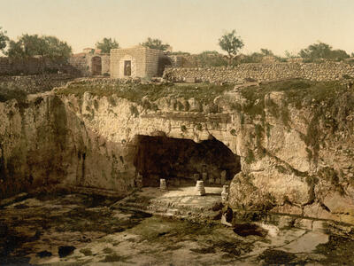 A large ancient rock-cut tomb with pillars and carvings set into a cliff, surrounded by grassy ground and stone walls, with small stone buildings and trees in the background.