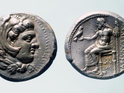 Front and back of an ancient silver coin. One side shows a profile of a man's head with curly hair, while the other side depicts a seated figure holding a bird, with Greek inscriptions beside the figure.