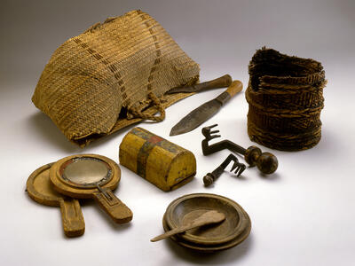 A collection of old household items including woven baskets, a wooden box, knives, keys, handheld mirrors, a wooden bowl with a spoon, and a wooden plate, arranged on a plain background.