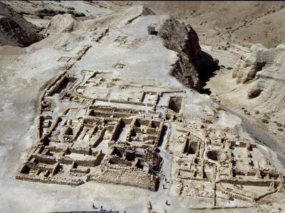Aerial view of ancient stone ruins in a desert landscape, showing rectangular foundations, walls, and pathways amid rocky terrain and sandy hills. People are visible exploring the site.
