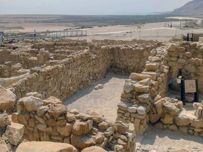 Ancient stone ruins with partially intact walls in a desert landscape. Distant visitors walk on a modern pathway under a hazy sky.