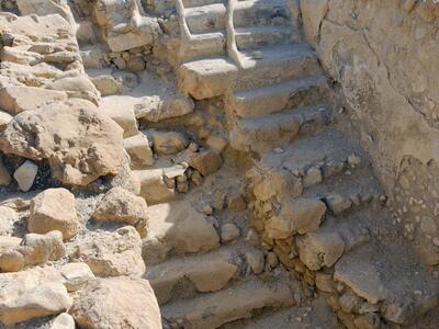 Ancient stone steps built into a rocky structure, surrounded by rough stone walls, with mountains visible in the background under a clear sky.
