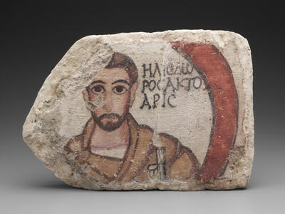Ancient clay block with painted plaster featuring a bearded man with large eyes, wearing a brown robe, next to Greek text and a red curved shape.