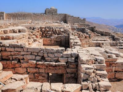 Ancient stone ruins atop a mountain, with multiple walls and chambers visible, overlooking a mountainous landscape under a clear sky.