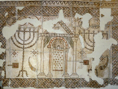 Ancient mosaic featuring a structure with pillars and an arch flanked by two menorahs, each flanked by a shofar and shovel, with intricate geometric borders and some damaged sections.